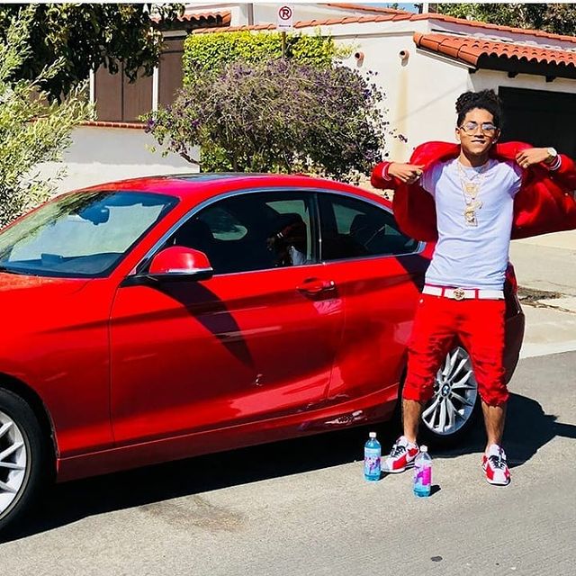 Jahking Guillory posing in a red suit and plain white t-shirt in front of his BMW 116.